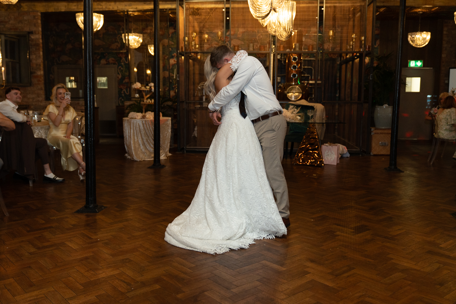 Couple sharing their first dance — embracing on a parquet floor beneath golden chandeliers, guests watching from the sides